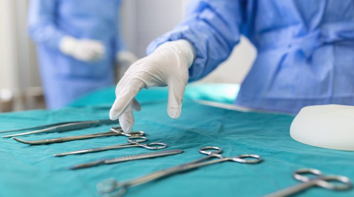 Nurse hand taking surgical instrument for group of surgeons at background operating patient in surgical theatre. Steel medical instruments ready to be used. Surgery and emergency concept